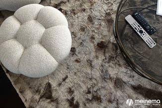 Witte ronde poef op grijze pluche vloerkleed, naast audioapparatuur en afstandsbedieningen op donker oppervlak.