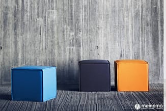 Drie kubische poefjes in blauw, zwart en oranje op gestreept grijs tapijt tegen houten wand.