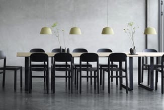 Vergadertafel met lichte houten blad en zwarte poten, omringd door zwarte stoelen. Vier hangende gele pendant lampen boven de