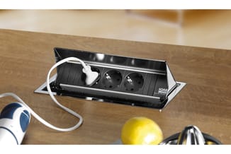 Zwart bureau-inbouwstroompaneel met twee stopcontacten en USB-poort op houten tafel, witte kabel aangesloten.
