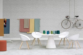 Witte ronde tafel met witte stoelen met houten poten, kleurrijke textielstalen aan muur, twee fietsen, grijze bakstenen wand.