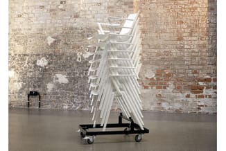 Stapelbare witte kunststof stoelen op zwart transportwagen tegen bakstenen muur in industriële ruimte.