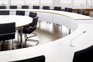 Moderne vergaderzaal met U-vormige witte vergadertafel, zwarte kantoorstoel en meerdere zwarte stoelen, hoge ramen, houten ac