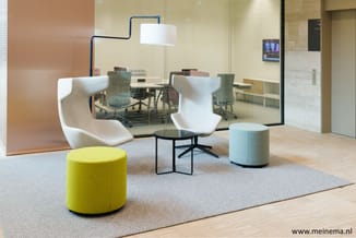 Moderne lounge-inrichting met gele poef, witte fauteuils, grijs poefje, zwarte tafeltje en blauwe hanglamp. Glazen wand naar