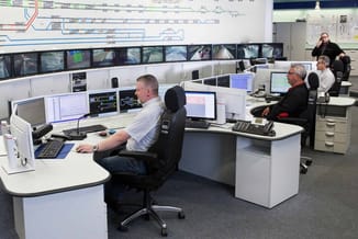 Controlecentrum met witte werkplekken, zwarte bureaustoelen, meerdere monitoren, schakelaars en technische apparatuur. Aan mu