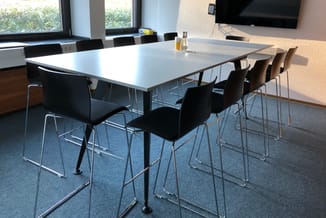 Moderne vergaderruimte met witte rechthoekige tafel, zwarte stoelen met chromen poten, TV aan wand, ramen met uitzicht.
