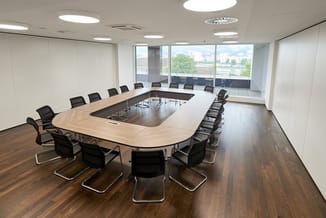 Vergaderkamer met halfronde houten vergadertafel, zwarte kantoorstoel, witte wanden, houten vloer en grote ramen met stadsuit