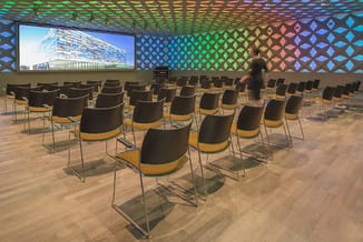 Presentatieruimte met rijen zwarte stoelen met gele zitting, houten vloer, gekleurde plafondpanelen en scherm met gebouw.
