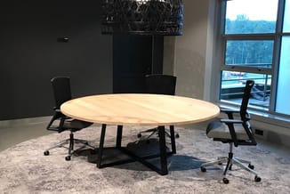 Ronde houten vergadertafel met zwart stalen onderstel in moderne kantoorruimte met zwarte bureaustoelen.