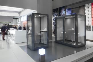Twee grijze belcellen met glazen wanden in een modern kantoorinterieur, met verlicht platform en kantoormeubelen op achtergro