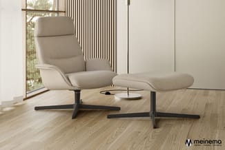 Beige relaxstoel met voetenbankje op zwart onderstel in minimalistisch interieur met houten vloer en raam.