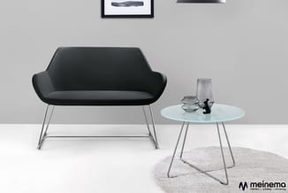 Zwarte moderne stoel met gebogende rugsteun op metalen poten naast glazen ronde tafel op grijs vloerkleed in lichte ruimte.