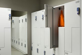 Rij witte kastjes met digitale sloten en nummering, één open kastje met oranje jas, tegen grijsgroene muur.