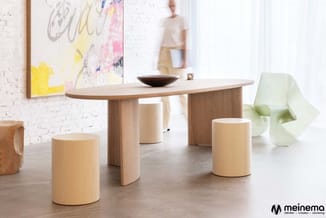 Houten ovale eettafel met cilindervormige houten krukken in natuurlijke tinten, in een lichte showroom met bakstenen muur en