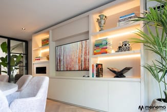 Moderne witte inbouwkast met verlichte open schappen, boeken, decoratie en kunstwerk. Grijze fauteuil en potplanten in contem