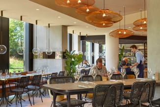 Modern restaurant met zwarte netwerk stoelen, houten tafels, koperen hangende lampen en grote ramen met uitzicht op groen.
