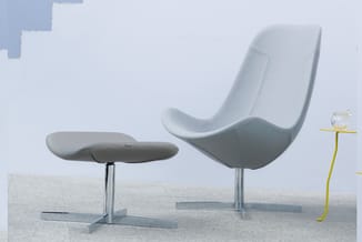 Grijze moderne loungeset met gebogen stoel, voetenbankje en geel bijzettafeltje op betonnen ondergrond tegen witte wand.