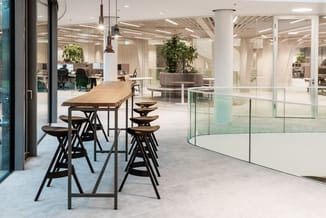 Modern kantoor met houten bartafel, zwarte barkrukken, glazen wanden, hanglamp, kolommen en groene plantenbakken.