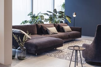 Bruine hoekbank met chaise longue, grijze kussens, in moderne kantoorruimte met groene planten, ronde bijzettafel en fauteuil