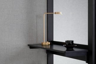 Zwart bureau met goudkleurige bureaulamp en zwart decoratief object, tegen grijze wand met zwarte stalen frame.