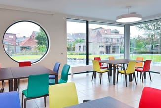 Moderne kantoorruimte met gekleurde stoelen (rood, geel, groen, blauw) rond zwarte tafels, grote ronde en rechthoekige ramen,