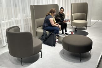 Moderne kantoorlounge met grijze ronde poef, gestoffeerde fauteuil en hoekbank met hoge rugleuning. Twee personen in gesprek