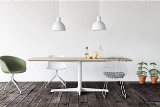 Houten eettafel met witte centrale voet, bediend door groene en witte stoelen, witte draadstoel, twee witte hanglampem en pot