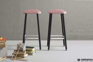 Twee roze barkrukken met zwarte poten en voetsteun, naast gestapelde boeken en een houten ladder op witte tafel tegen grijze