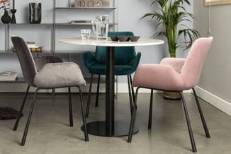 Ronde witte eettafel met zwart onderstel, omgeven door drie stoelen in beige, groen en roze fluweel met metalen poten. Achter
