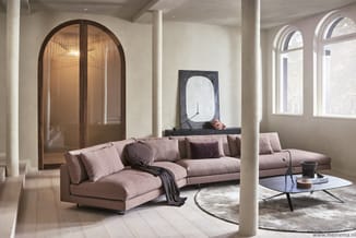 Moderne woonkamer met roze-beige hoekbank, witte zuilen, ronde salontafel, archiefkast en grote raampartijen.