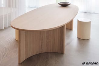 Lichte houten ovale vergadertafel met gebogen voet naast cylinder bijzettafel in minimalistische kantooromgeving.