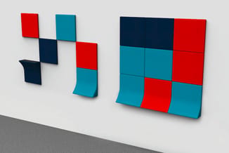 Modulaire wandpanelen in rood, petrol en donkerblauw, volgens verschillende configuraties tegen witte wand opgesteld.