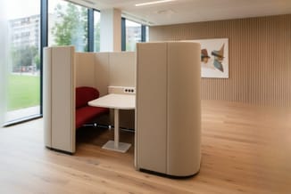 Akoestische belcel met beige stoffering, rode stoel en wit tafelblad in modern kantoor met houten vloer.