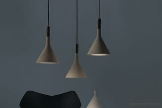 Drie betonnen hangende lampen in grijs/taupe met zwarte ophanging, tegen grijze wand, met zitmeubel onderaan.