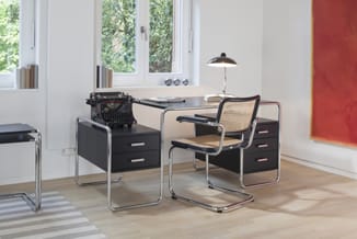 Kantoorwerkplek met zwart bureau met laden, rieten stoel, typemachine, bureaulamp, houten vloer, grote ramen en rode muurpane