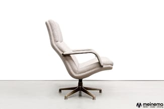 Beige/crème gestoffeerde draaibare fauteuil met hoge rug, armleuningen en bruin metalen voetenkruis op grijs oppervlak.