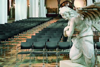 Zwarte kantoorstoel in een kerkzaal met rijen stoelen en klassieke zuilen, naast een wit stenen engelenbeeld.