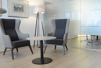 Moderne vergaderhoek met twee grijze oorfauteuils, zwart rond tafeltje, zwarte staande lamp en glazen wand in lichte kantoorr