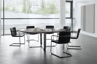 Ronde zwarte vergadertafel met zes zwarte kantooorstoelen met metalen frames in moderne kantoorruimte met grote ramen.