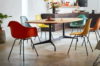 Ovale vergadertafel met zwart onderstel en houten blad, omgeven door gekleurde stoelen in rood, geel, groen en bruin met zwar
