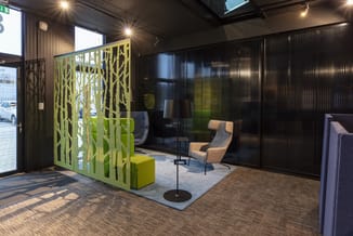 Showroom met groen decoratief scheidingspaneel, beige fauteuil, zwarte verticale lamellen, grijze loungebank en houten vloer.