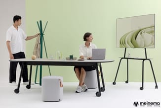 Moderne werkplek met zwarte vergadertafel op wielen, witte pouf, man in wit overhemd bij groene kapstok, vrouw aan laptop, be