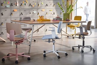 Modern kantoor met houten tafel, gekleurde bureaustoelen (roze, lichtblauw, geel, grijs), grijze kasten met notities aan muur