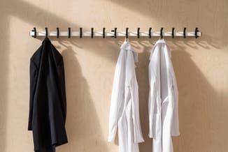 Witte kapstok met zwarte haakjes aan beige muur, waaraan een zwart kledingstuk en twee witte overhemden hangen.