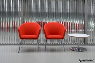 Twee rode stoelen met metalen poten en een ronde witte tafel in een moderne kantoorruimte met verticale jaloeziën.