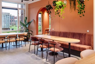Moderne kantoorruimte met terracotta wand, hoekbank in roestbruin, houten eettafel, fluwelen stoelen en hangplanten.