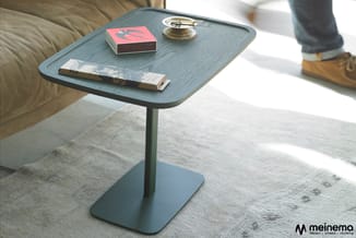 Staande bijzettafel met blauwe houten tafelblad en metalen voet, tegen beige bank, met boeken en decoratie erop.
