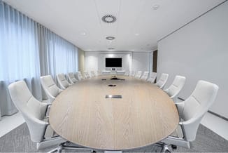 Moderne vergaderruimte met ovale houten tafel, grijze stoelen, witte wanden, gordijnen en display aan wand.