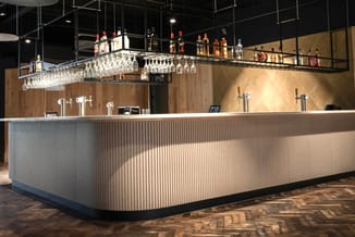Moderne bartafel met geribbeld lichtgrijs front en zwarte sokkel. Daarachter houten wandafwerking, hangend glaswerk en flesse