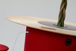 Houten vergadertafel met rode stoffen zijpaneel, grijs blad, groene plant en witte stroomkabel, tegen grijze achtergrond.